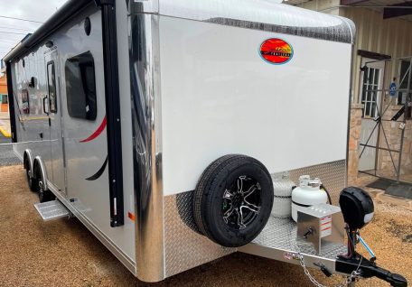 A sleek, silver travel trailer with black wheels, a propane tank, and a prominent logo, parked on gravel near a building entrance.