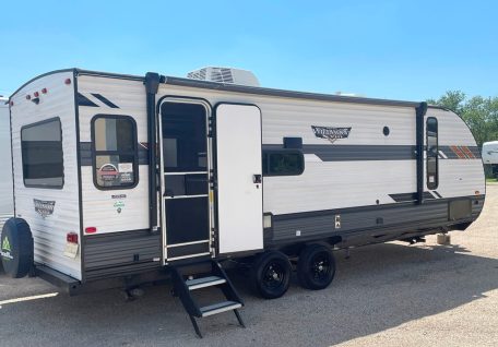 A side view of a modern white and gray travel trailer with a door, steps, and windows, parked outdoors under a clear blue sky.