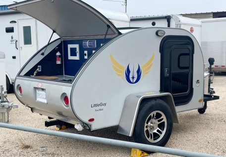 A compact silver teardrop trailer with a raised hatch revealing interior features, adorned with a blue and gold emblem.