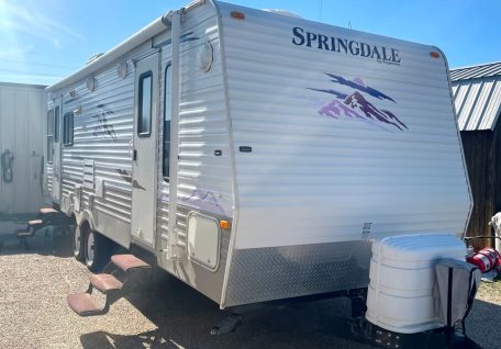 A white Springdale travel trailer parked in gravel, featuring mountain graphics, steps, and a propane tank on the front.