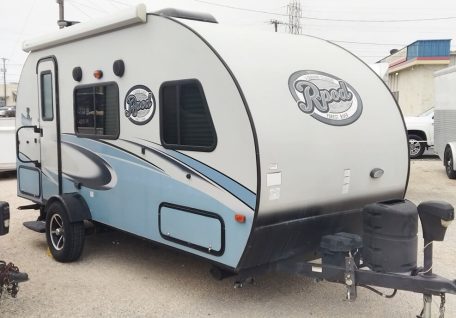 A compact, light blue and white RPod travel trailer parked on a gravel lot, featuring a rounded top and large windows.