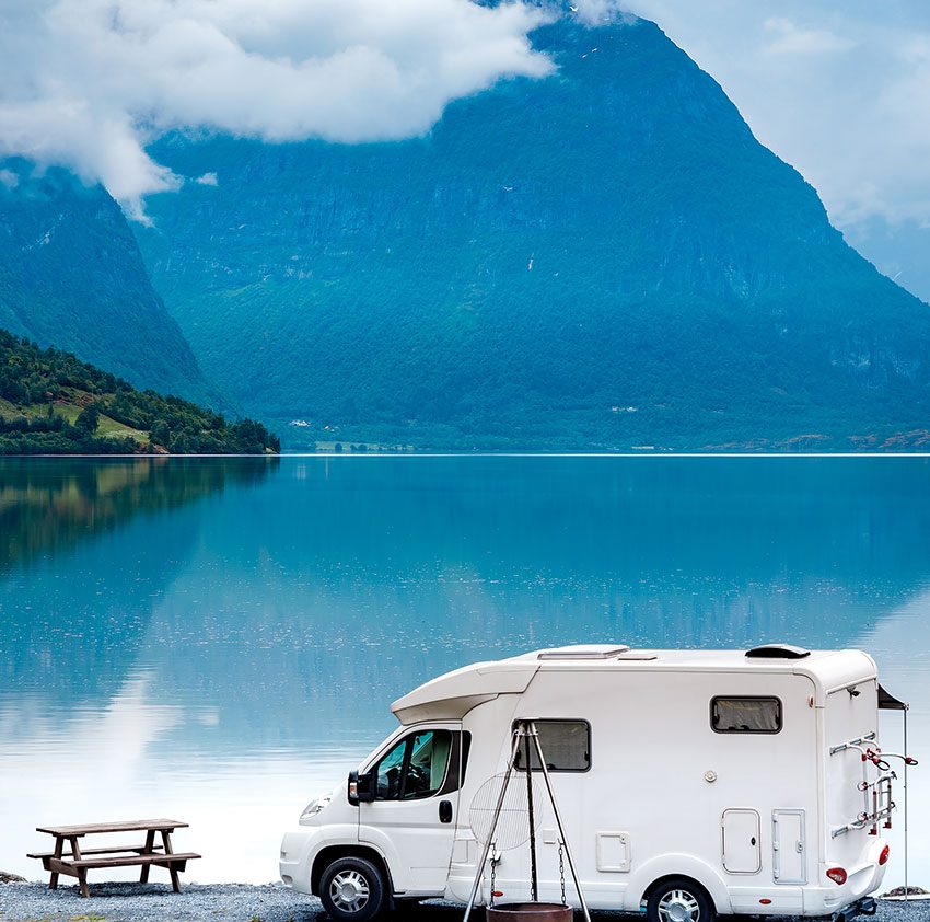 A white motorhome parked by a serene lake, surrounded by green mountains and cloudy skies, with a picnic table nearby.