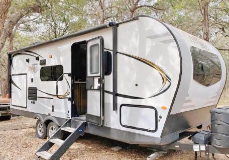 A sleek, modern travel trailer parked in a wooded area, featuring a metal staircase leading to the entrance and large windows.