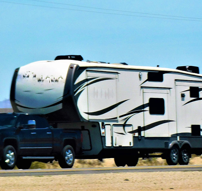 A black pickup truck towing a sleek, white fifth-wheel RV with black accents, set against a clear blue sky and arid landscape.