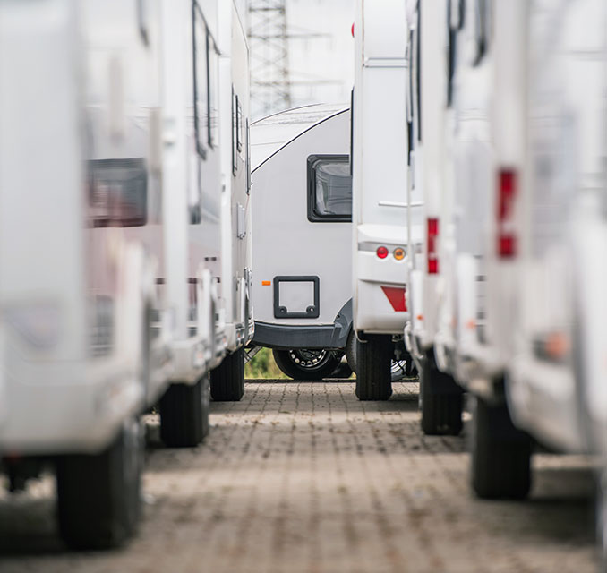 A narrow pathway between rows of white travel trailers, with one gray trailer visible in the background, surrounded by greenery.